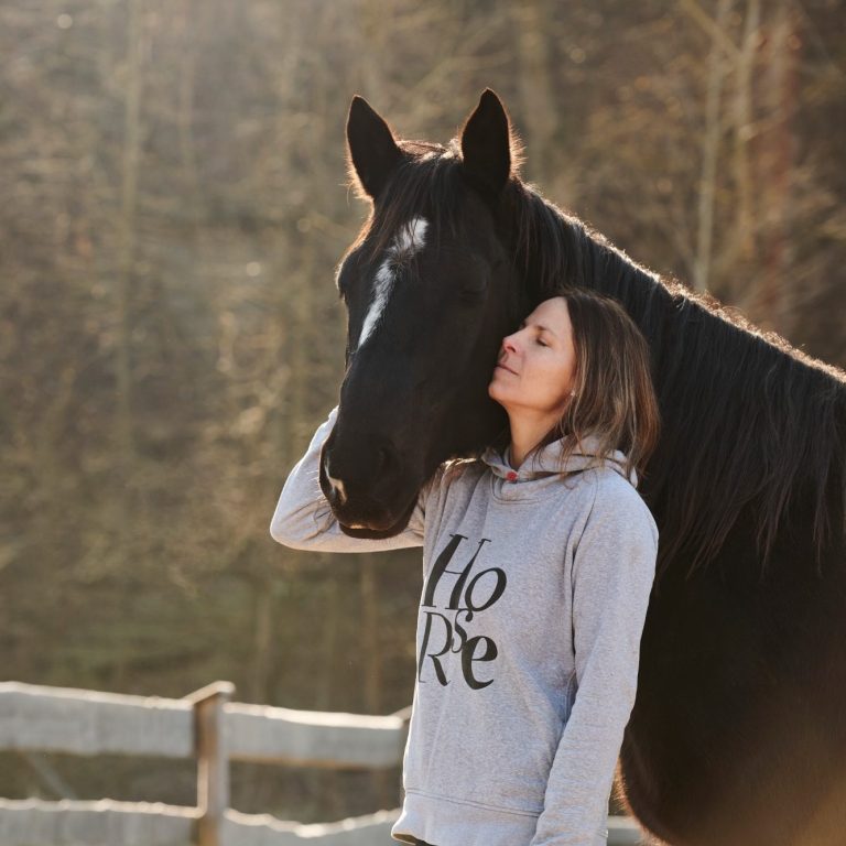 Frau umarmt ein schwarzes Pferd in einer sonnigen, ländlichen Umgebung.