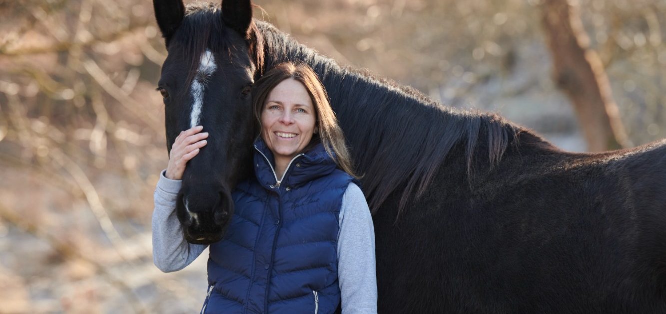 Frau mit dunklem Pferd in ländlicher Umgebung, lächelnd und freundlich.