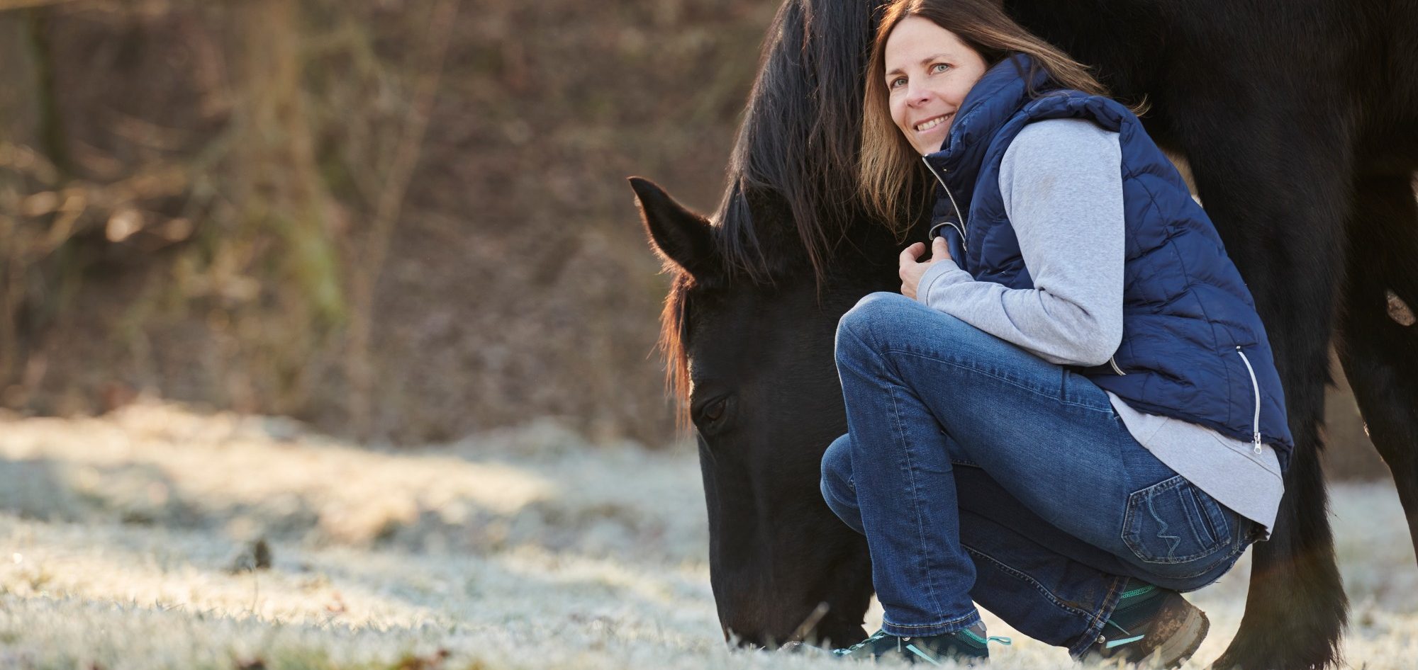 Horsebrain Coaching Denise Wiegand Frau in Jeans und Weste kniet neben einem Pferd in der Natur.
