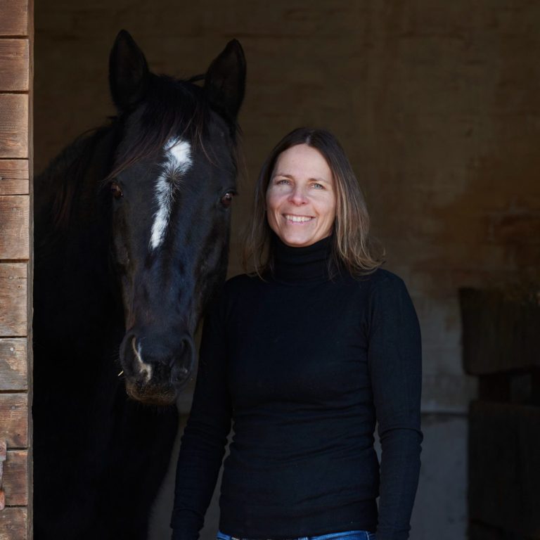 Horsebrain Coaching Denise Wiegand Frau mit Langhaar in schwarzem Pullover steht neben einem schwarzen Pferd.