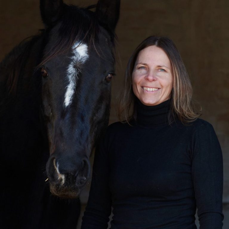 Denise Weigand in schwarzem Rollkragenpullover steht neben einem schwarzen Pferd mit weißen Streifen auf dem Kopf.